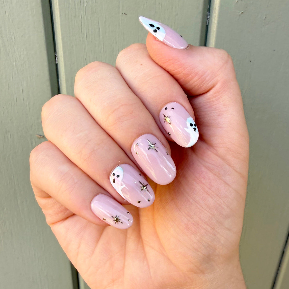 A close-up of a hand with round shaped nails painted in a light nude base with white chrome ghosts and gold star details, set against a muted green wooden background.