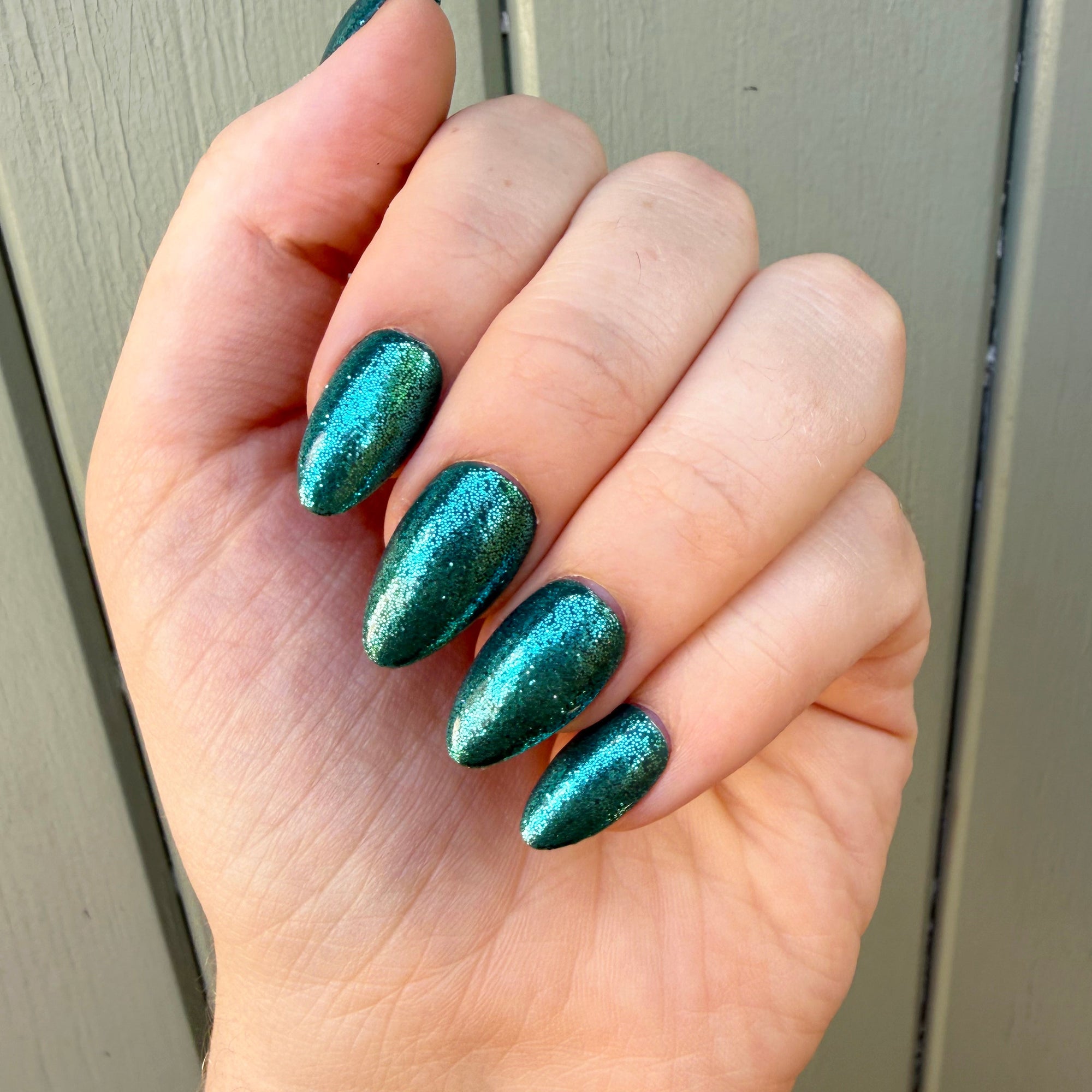 A close-up of a hand with almond-shaped nails painted in a shimmering metallic teal green nail polish, set against a neutral gray paneled background.